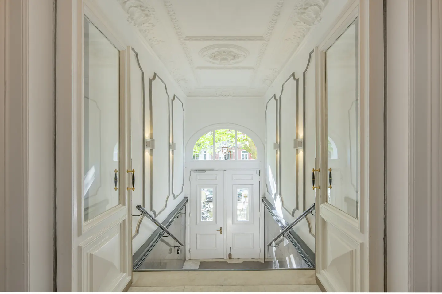 Elegant white hallway with ornate ceiling molding, mirrored walls, and a staircase leading to a glass-paneled front door.