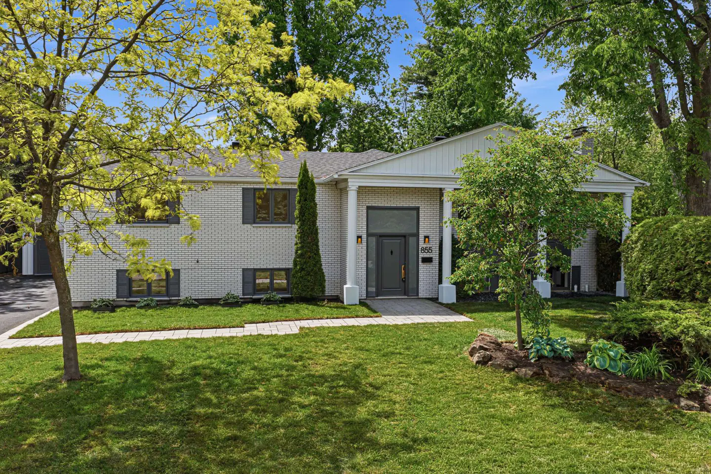 Single-story white brick house with a dark gray front door, white columns, and a lush green lawn under a blue sky.