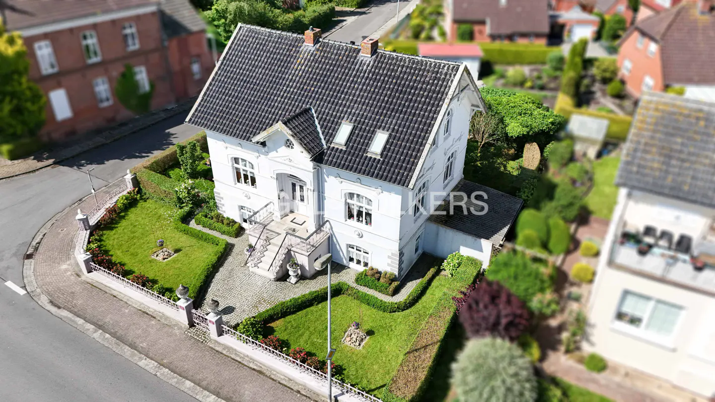 Aerial view of a white two-story house with a dark gray roof, surrounded by green lawns and hedges.