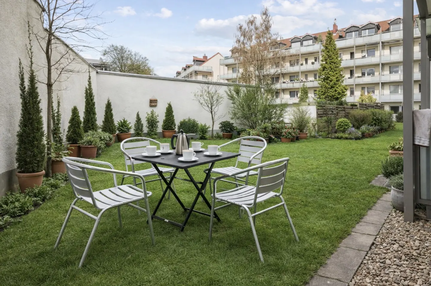 A black table with four white chairs sits on a green lawn, set for tea with a silver pot and white cups. A white wall and trees surround the yard.