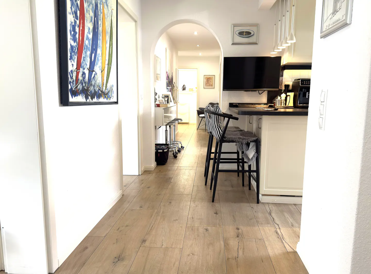 Interior view of a hallway with light wood floors, leading to a kitchen with black and white bar stools. A colorful painting hangs on the left wall.