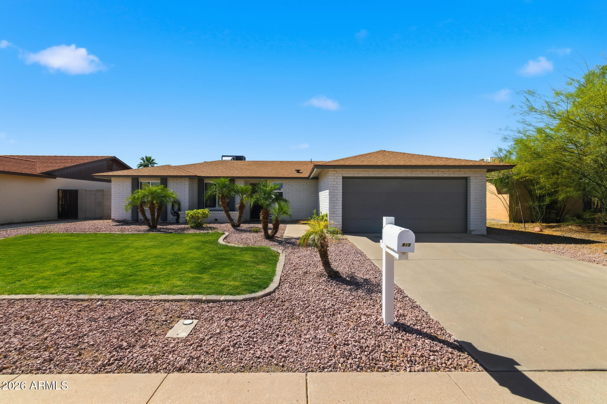 A single-story white brick house with a brown roof, a gray garage door, and a green lawn. Palm trees and a white mailbox are in the front yard.