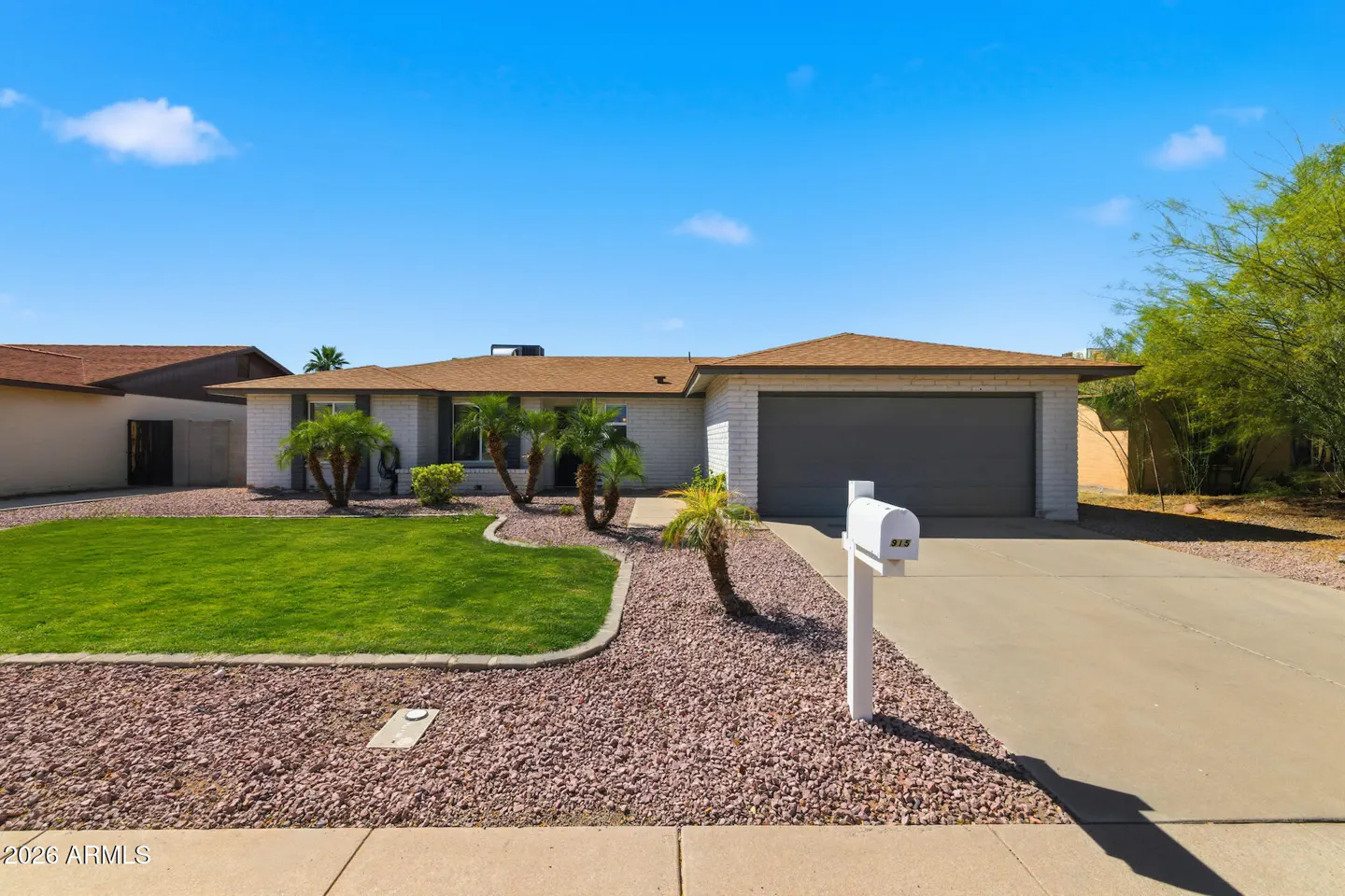 A single-story white brick house with a brown roof, a gray garage door, and a green lawn. Palm trees and a white mailbox are in the front yard.