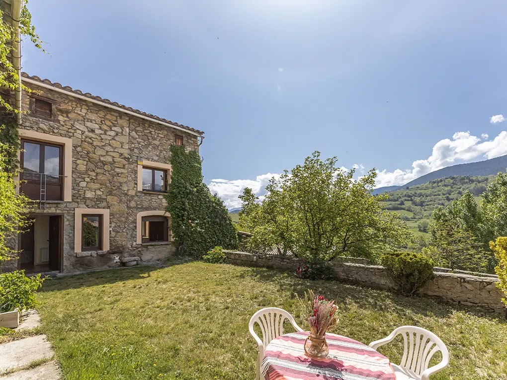 Stone house with ivy, lawn, and outdoor table with flowers. Mountains and blue sky in background.
