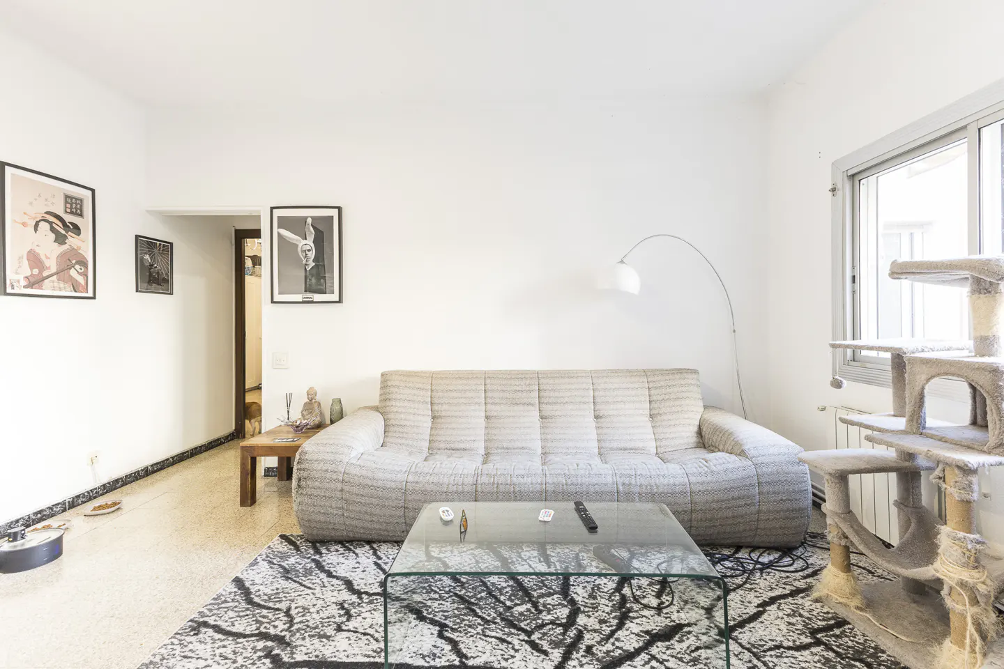 Bright living room with a beige sofa, glass table, and a black and white rug. A cat tree stands near a window. Artwork adorns the white walls.