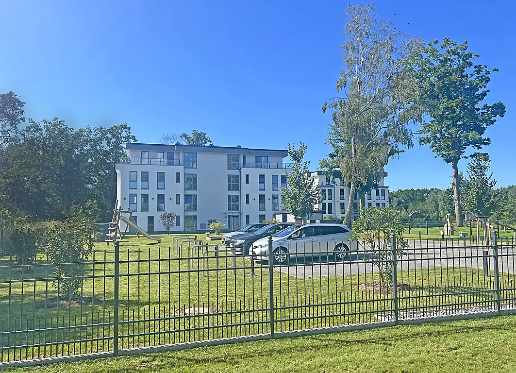 Modern white apartment building with balconies, cars parked in front, playground, and black metal fence on a sunny day.