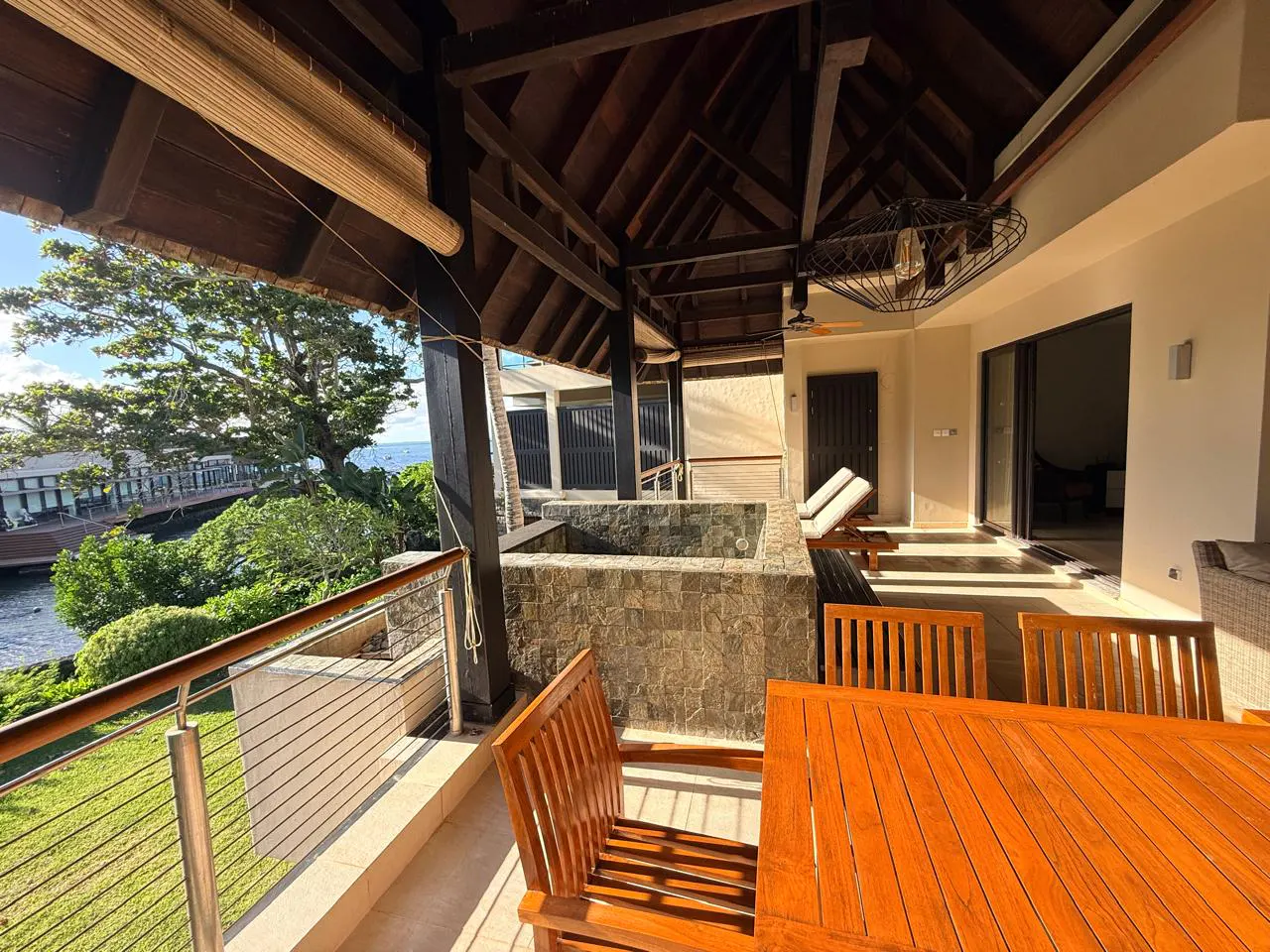 Covered patio with a stone hot tub, wooden table and chairs, and lounge chairs. Ocean view with green trees and a building in the background.