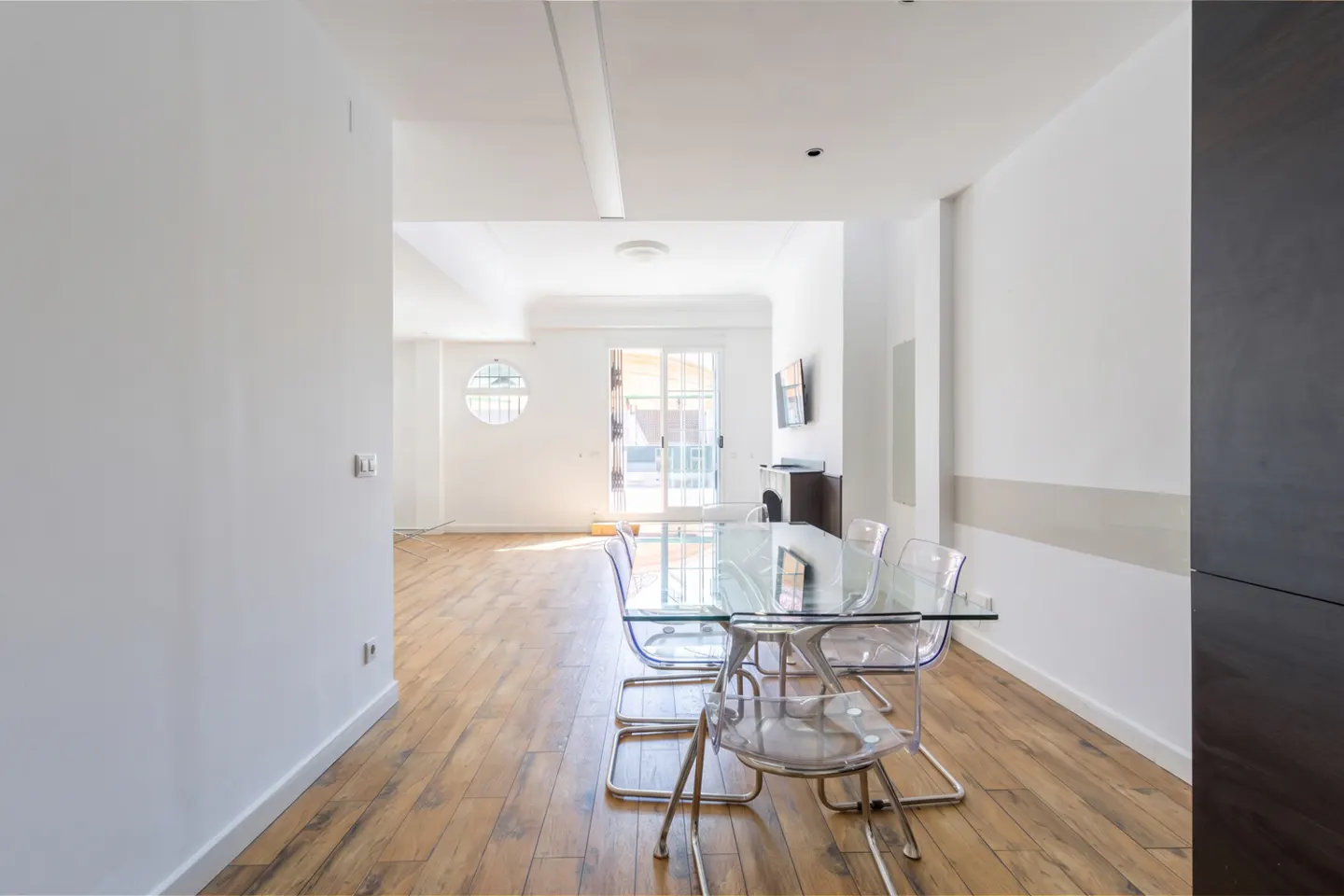 Bright, modern dining area with a glass table and clear chairs on a wood floor. White walls and natural light fill the space.