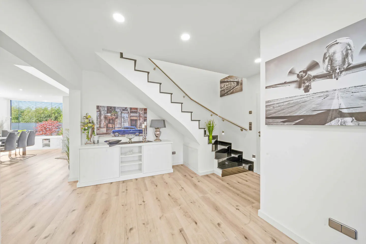Bright, modern home interior with light wood floors, white walls, and a staircase with black steps. Artwork and decor add visual interest.