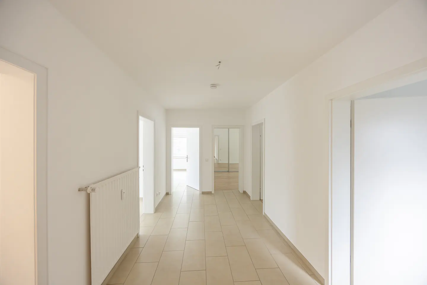 A bright, empty hallway with white walls, light-colored tile flooring, and several open doorways leading to other rooms.