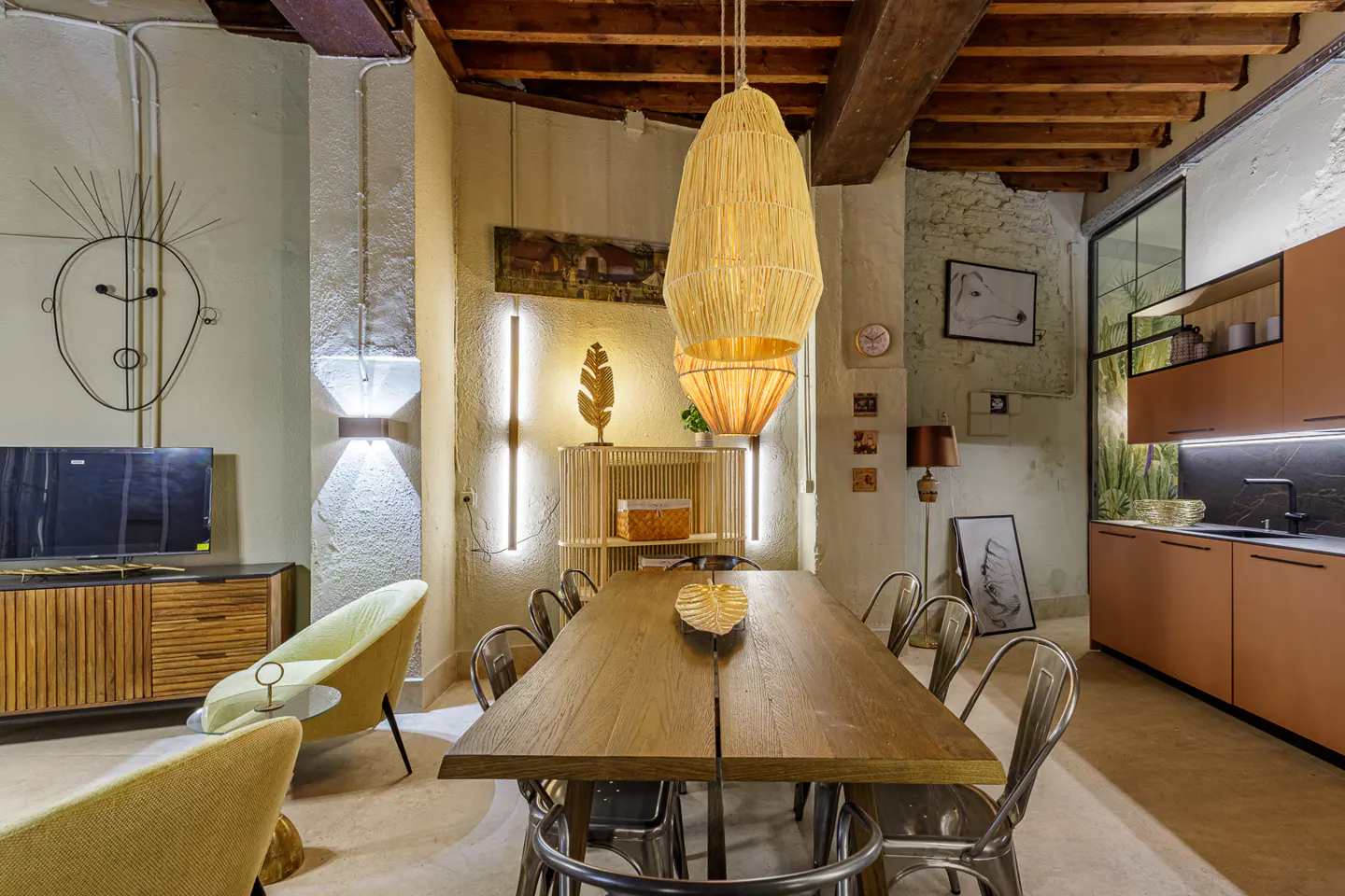 A modern dining room with a long wooden table, metal chairs, and a straw pendant light. The room has exposed beams and a kitchen area in the background.
