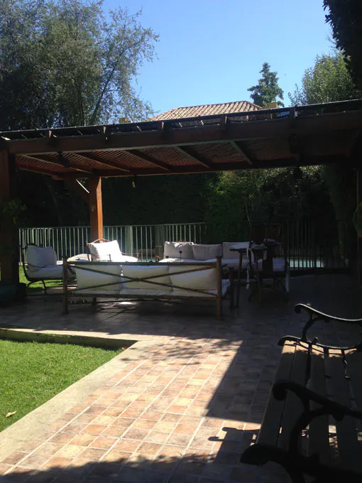 Outdoor patio with a wooden pergola, white cushioned seating, and brick flooring. Green lawn and trees in the background.
