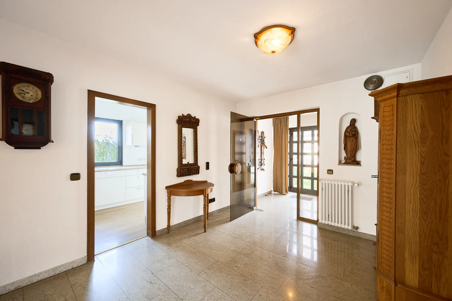 A bright foyer with a marble floor, white walls, and wooden accents. A clock hangs on the left, and a glass door leads to the outside.
