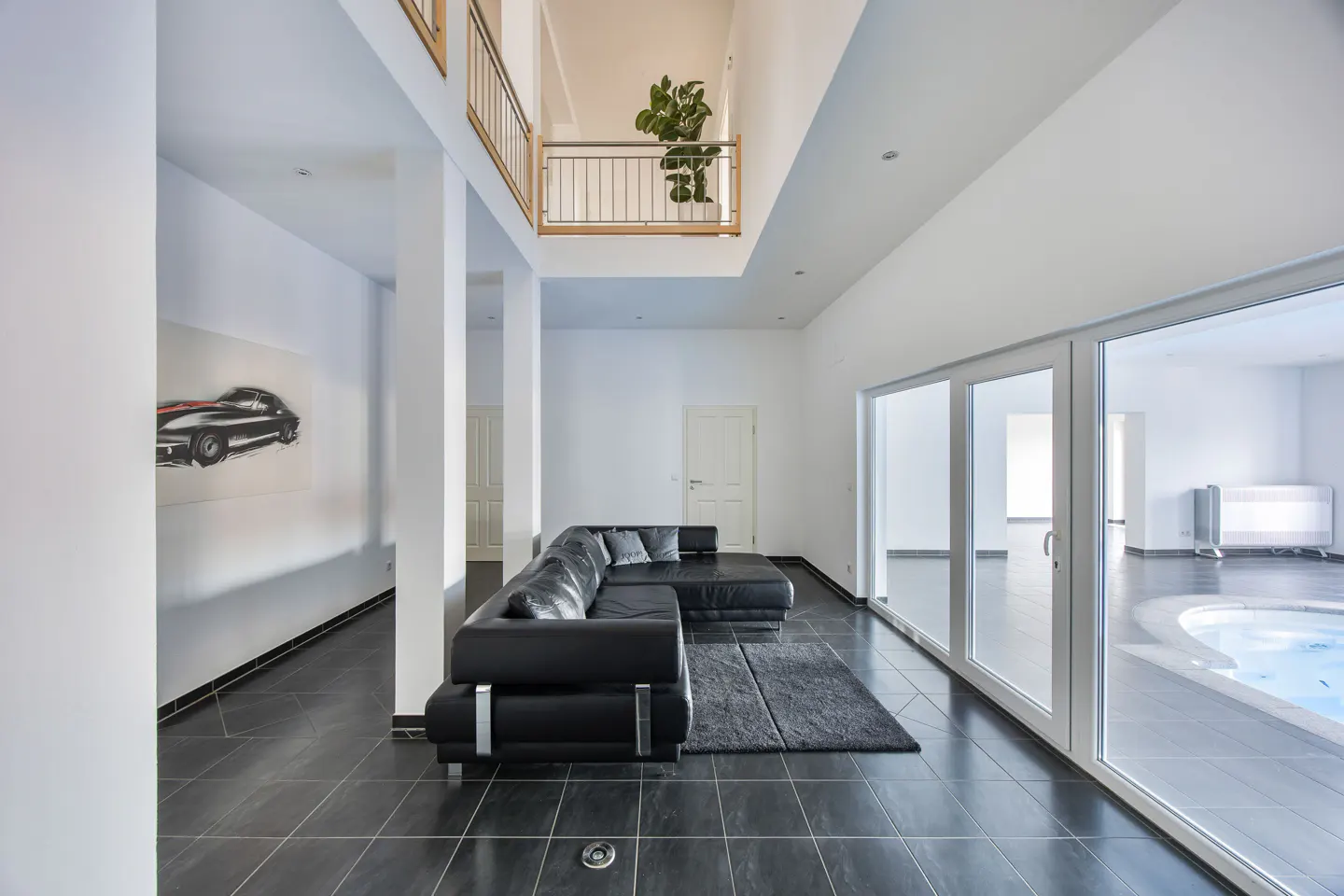 Bright, modern living room with black leather sofa, dark tile floor, and white walls. Balcony with plant overlooks the space. Hot tub visible through glass doors.