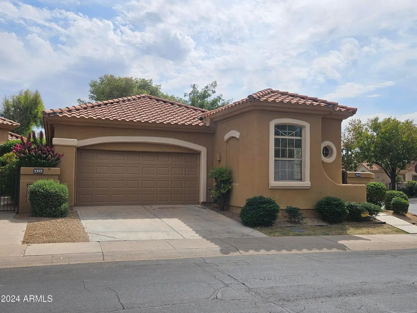 Tan stucco house with a brown garage door and a red tile roof under a cloudy sky.