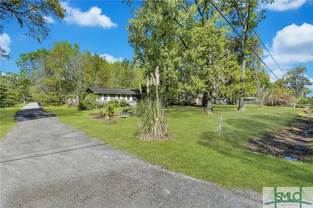 A single-story green house with a gray roof sits on a large grassy lot with trees and a gravel driveway.