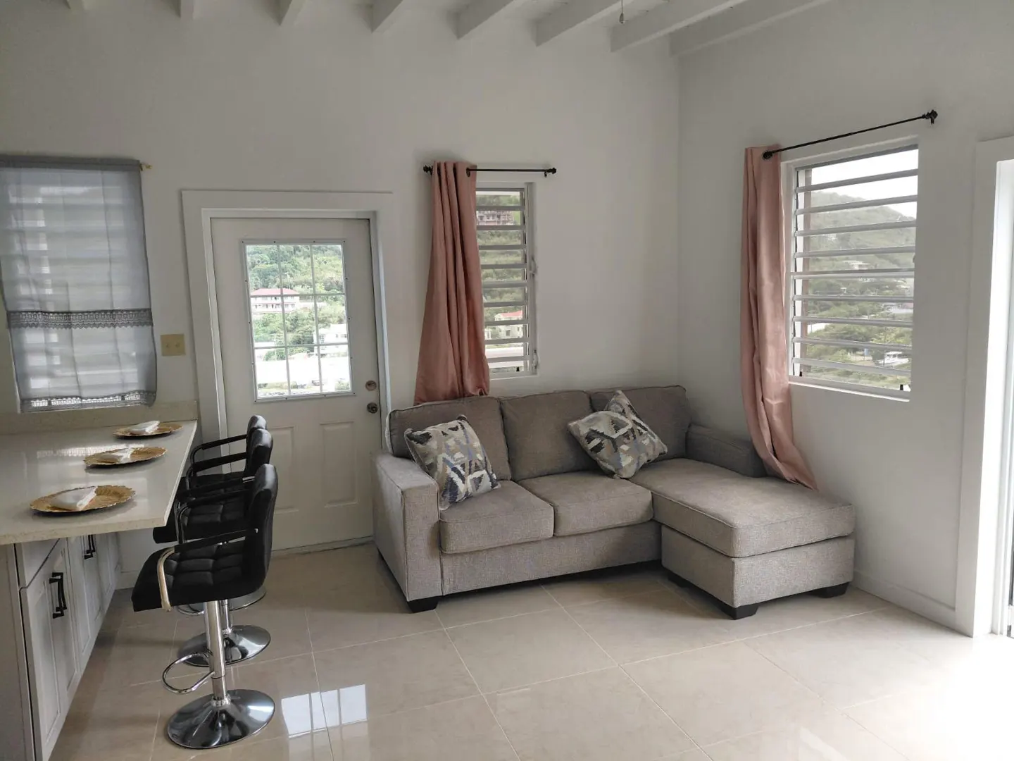 Bright living space with a gray sectional sofa, pink curtains, and a breakfast bar with black stools. View of greenery outside.