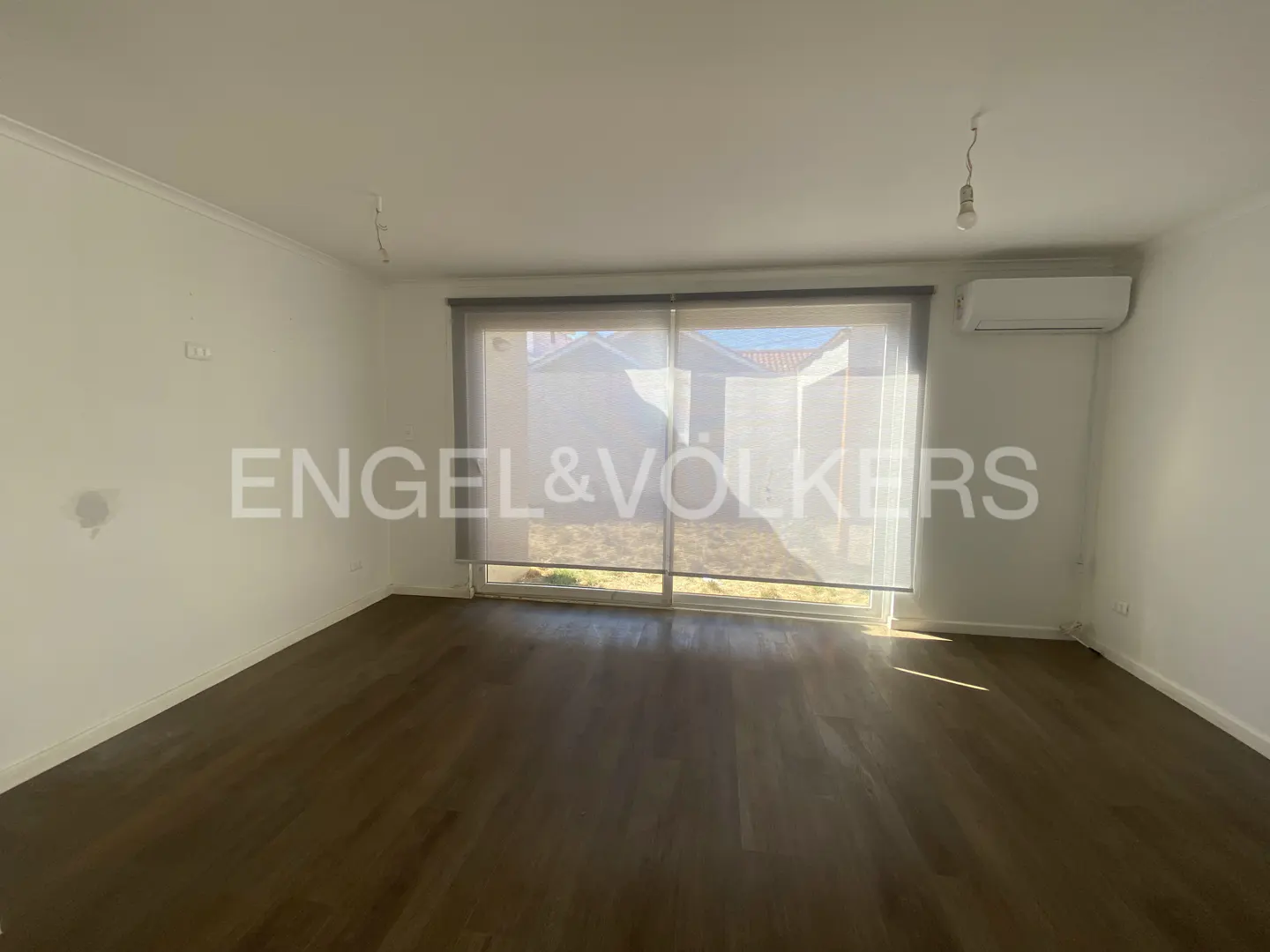 Bright, empty room with dark wood floors and white walls. Large window with sheer blinds overlooks a yard. Engel & Völkers logo is superimposed.