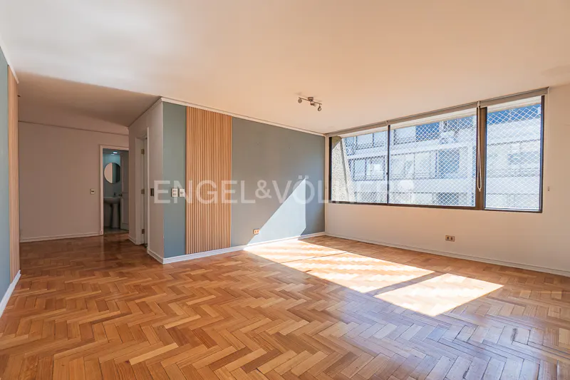 Bright, empty room with herringbone wood floors, large windows, and blue-gray walls. A bathroom is visible through a doorway.