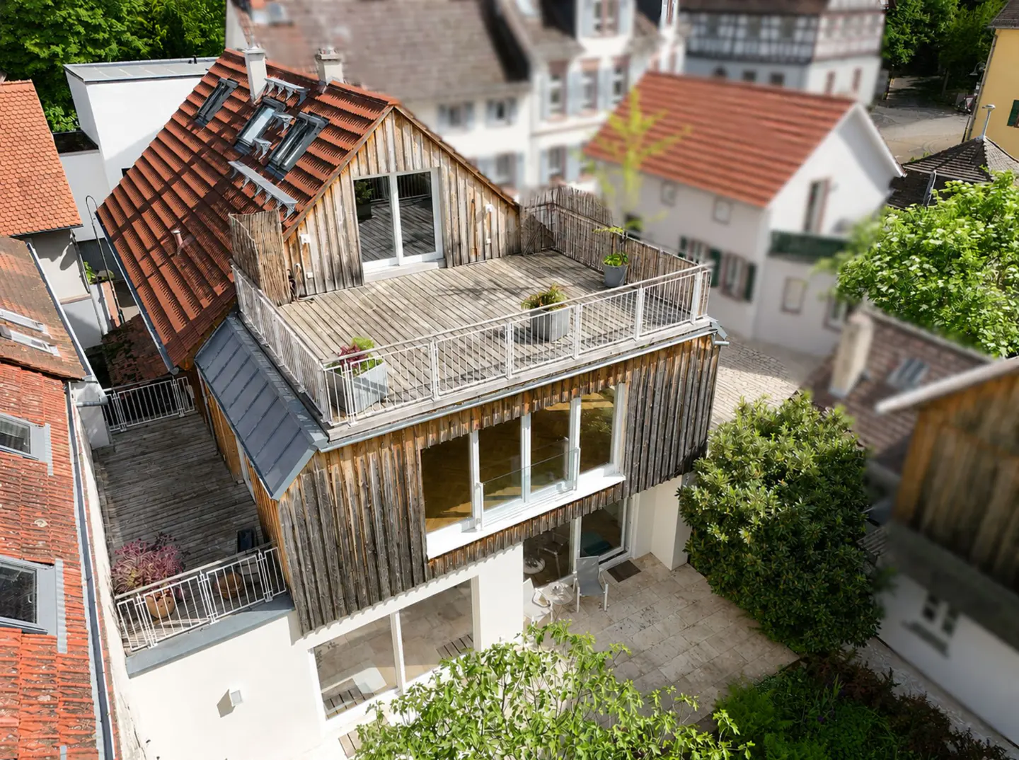 Aerial view of a modern house with a wooden facade and rooftop deck, surrounded by other houses and greenery.