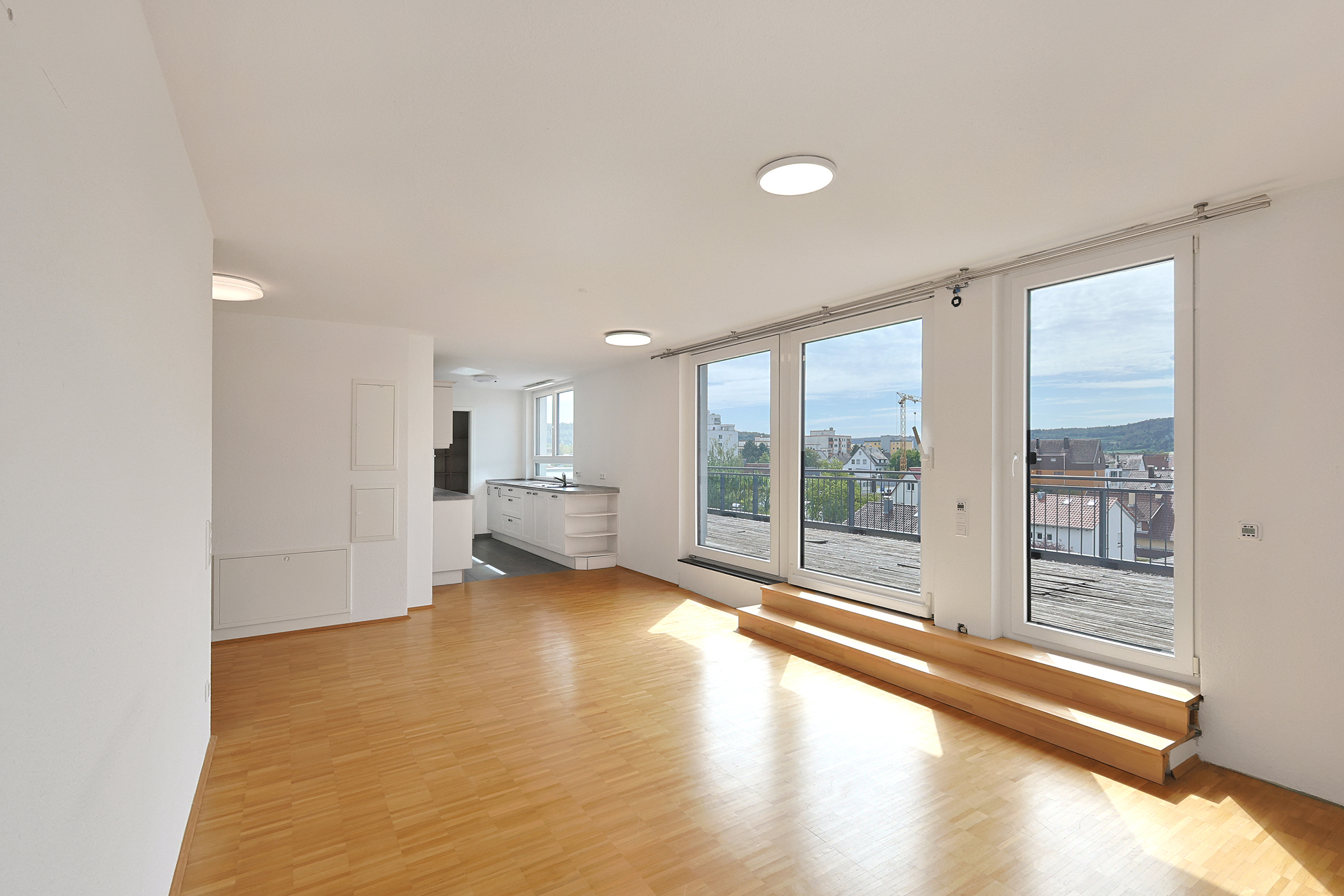 Bright, open-plan living area with light wood floors, white walls, and large windows leading to a balcony. [AI-generated text]