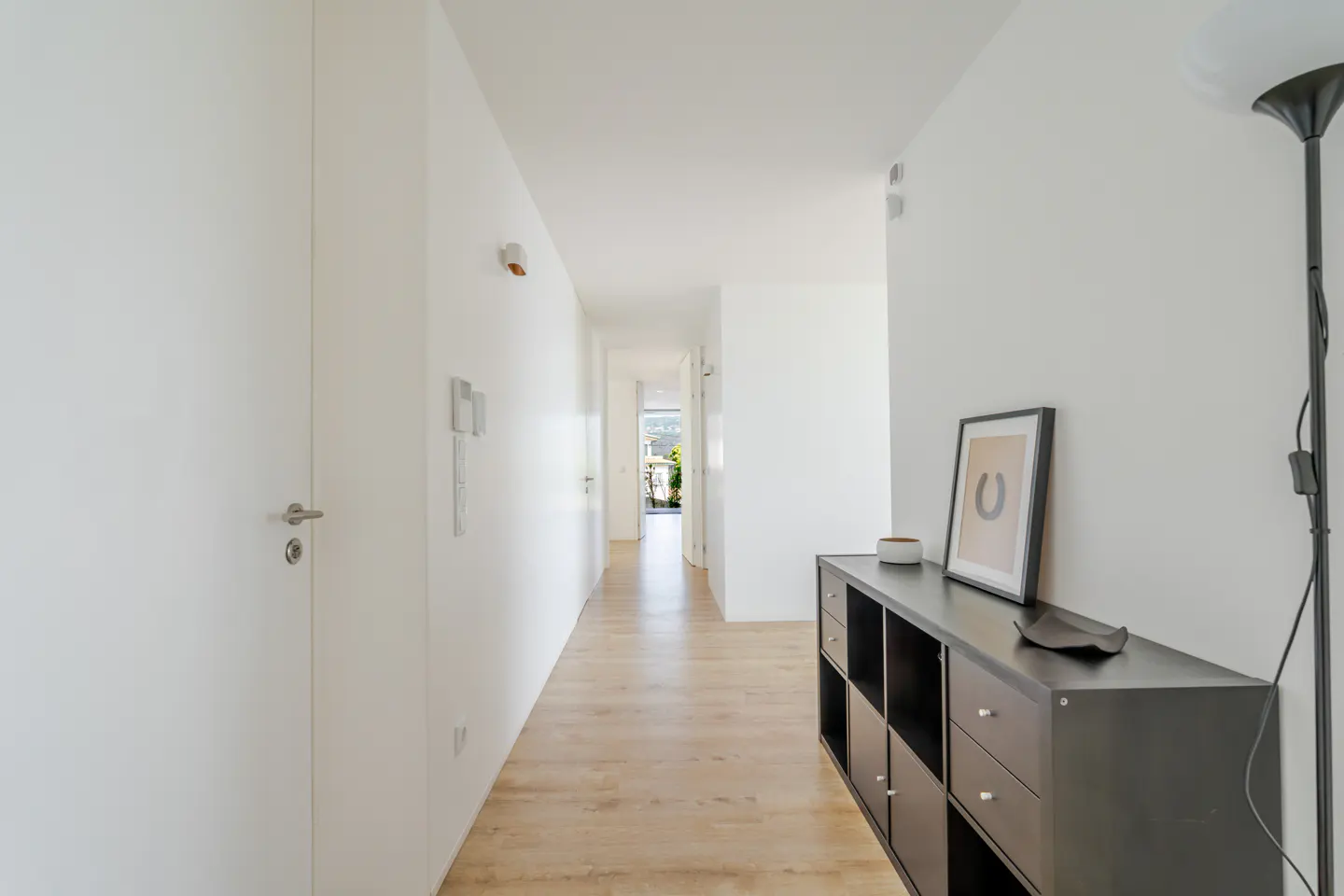 Bright hallway with light wood floors, white walls, a dark storage unit, and a floor lamp leading to a sunlit doorway.