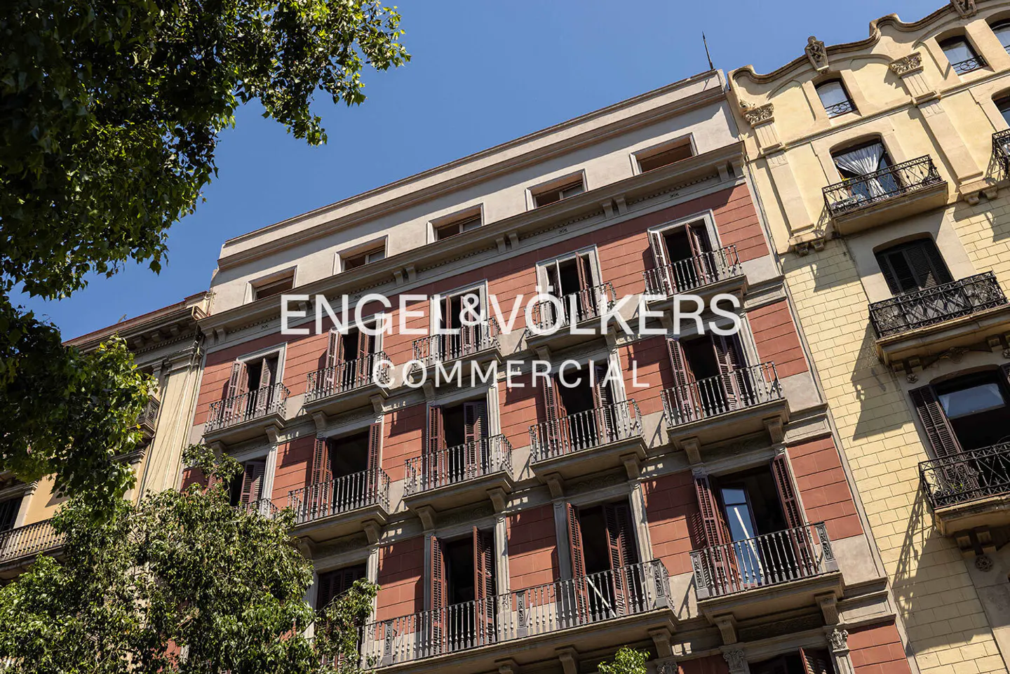 Exterior of a multi-story building with balconies, red facade, and open shutters under a blue sky. Trees are visible on the left.