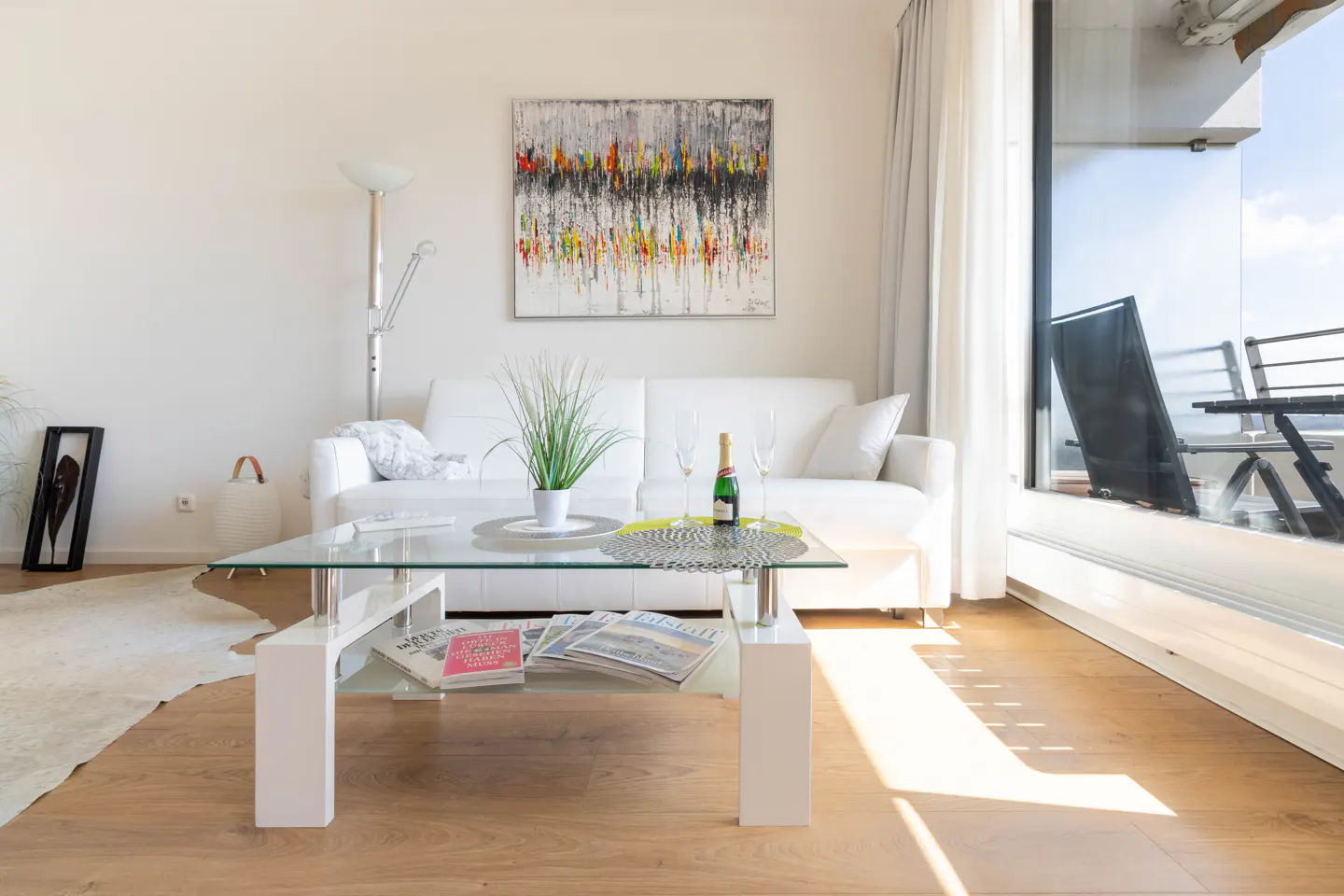 Bright living room with white sofa, glass table, and balcony. Champagne bottle and glasses sit on the table. Abstract art hangs on the wall.