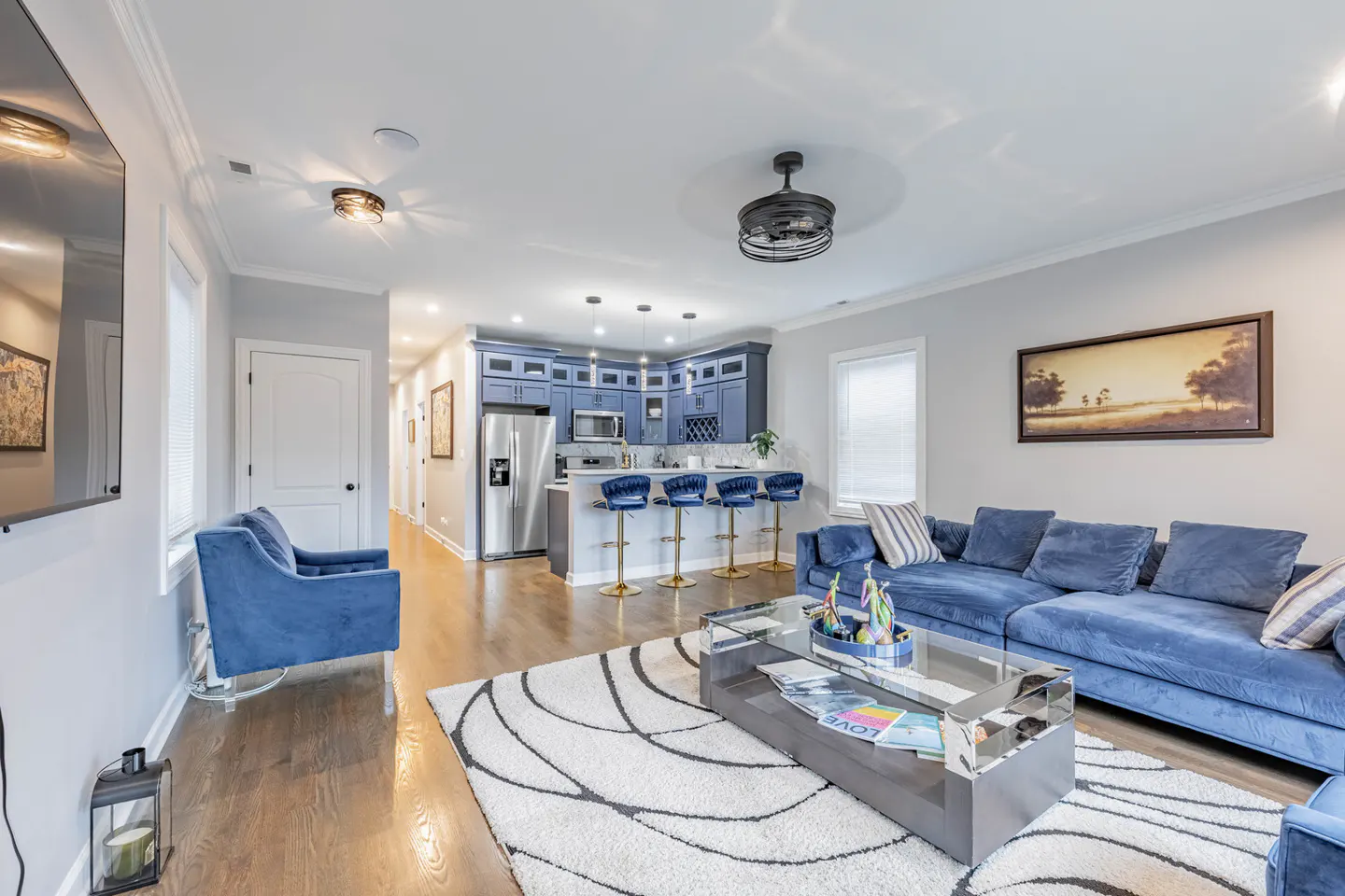 Open-concept living room with blue velvet furniture, a white rug, and a kitchen with blue cabinets and stainless steel appliances.