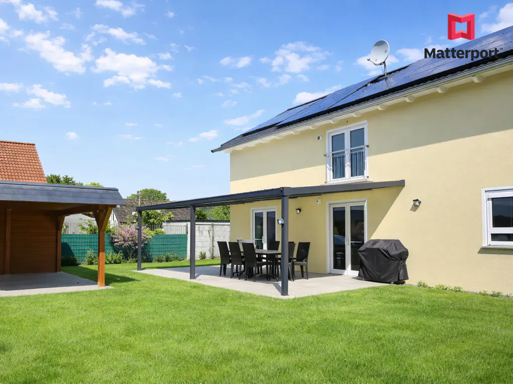 Backyard view of a two-story yellow house with solar panels, a patio with a table and chairs, a grill, and a wooden carport.