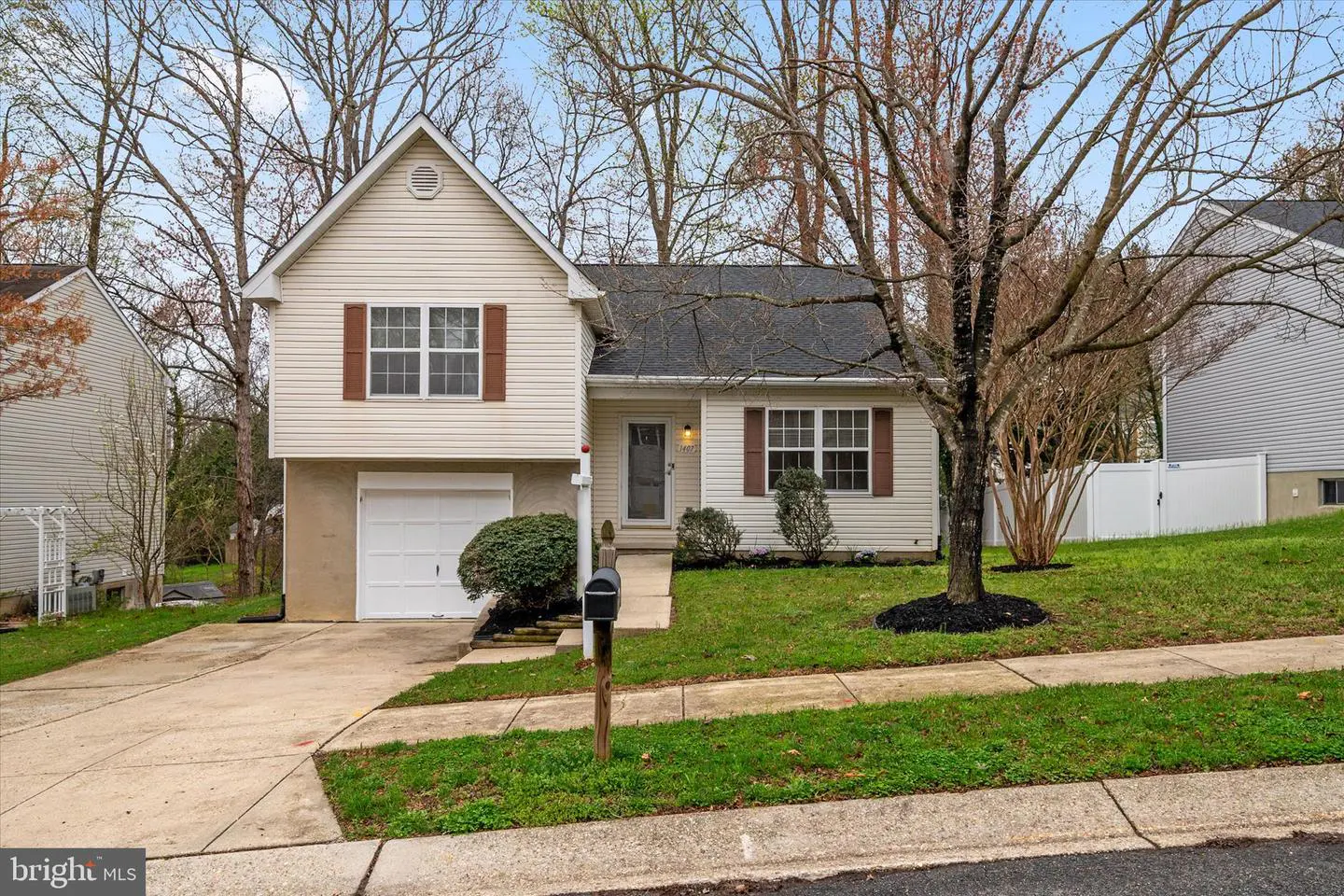 Two-story beige house with brown shutters, a white garage door, and a front yard with a tree and mailbox.