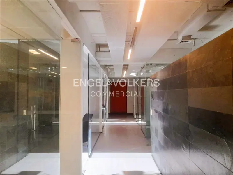 Office hallway with glass-walled offices, white ceiling with lights, and a red door at the end. "Engel & Volkers Commercial" logo visible.