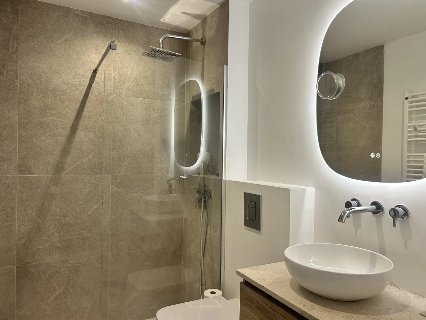 Modern bathroom with a walk-in shower, beige marble tiles, a white oval sink, and a large illuminated mirror.