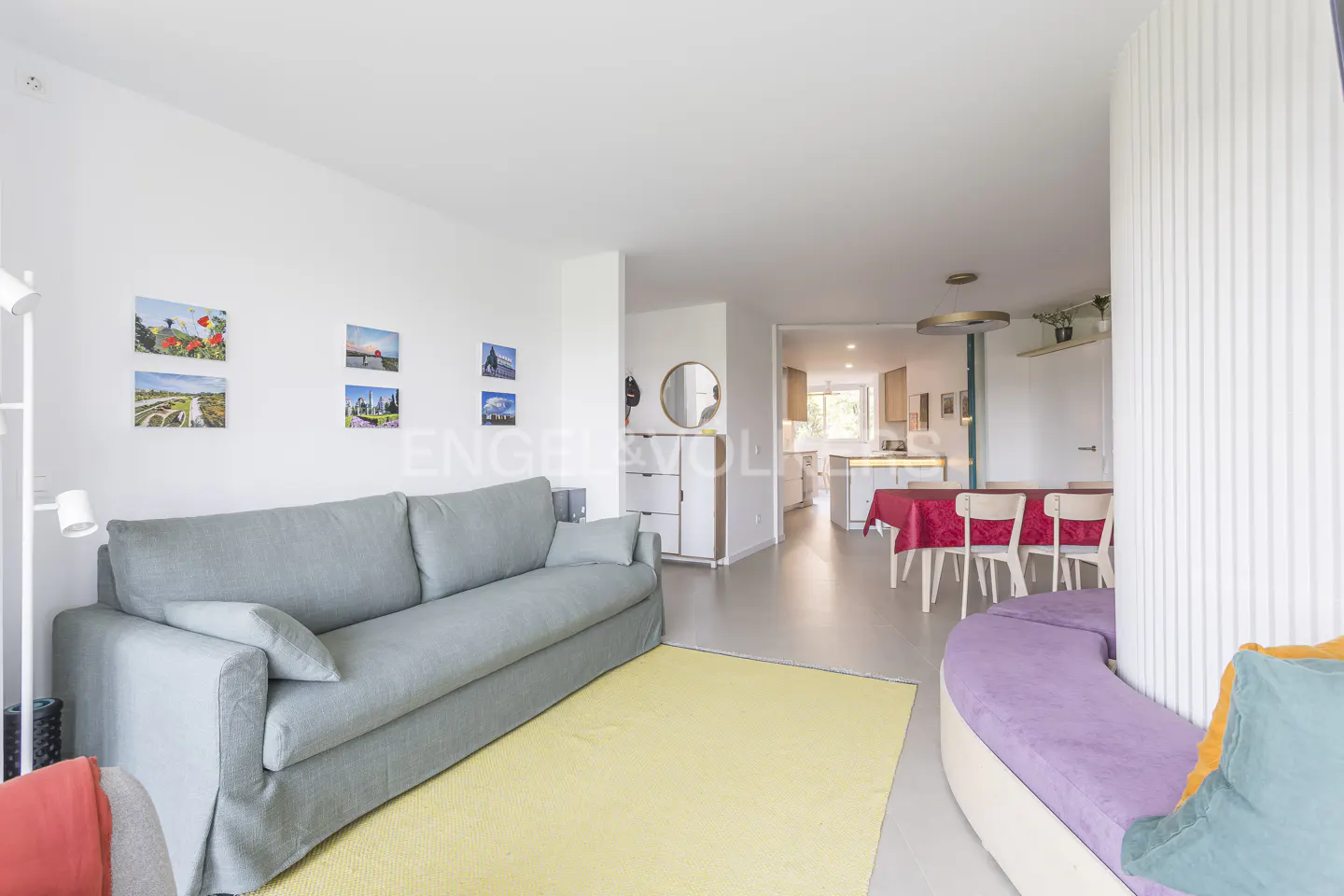 Open-concept living space with a gray sofa, yellow rug, and a dining area with a red tablecloth. Kitchen in the background.