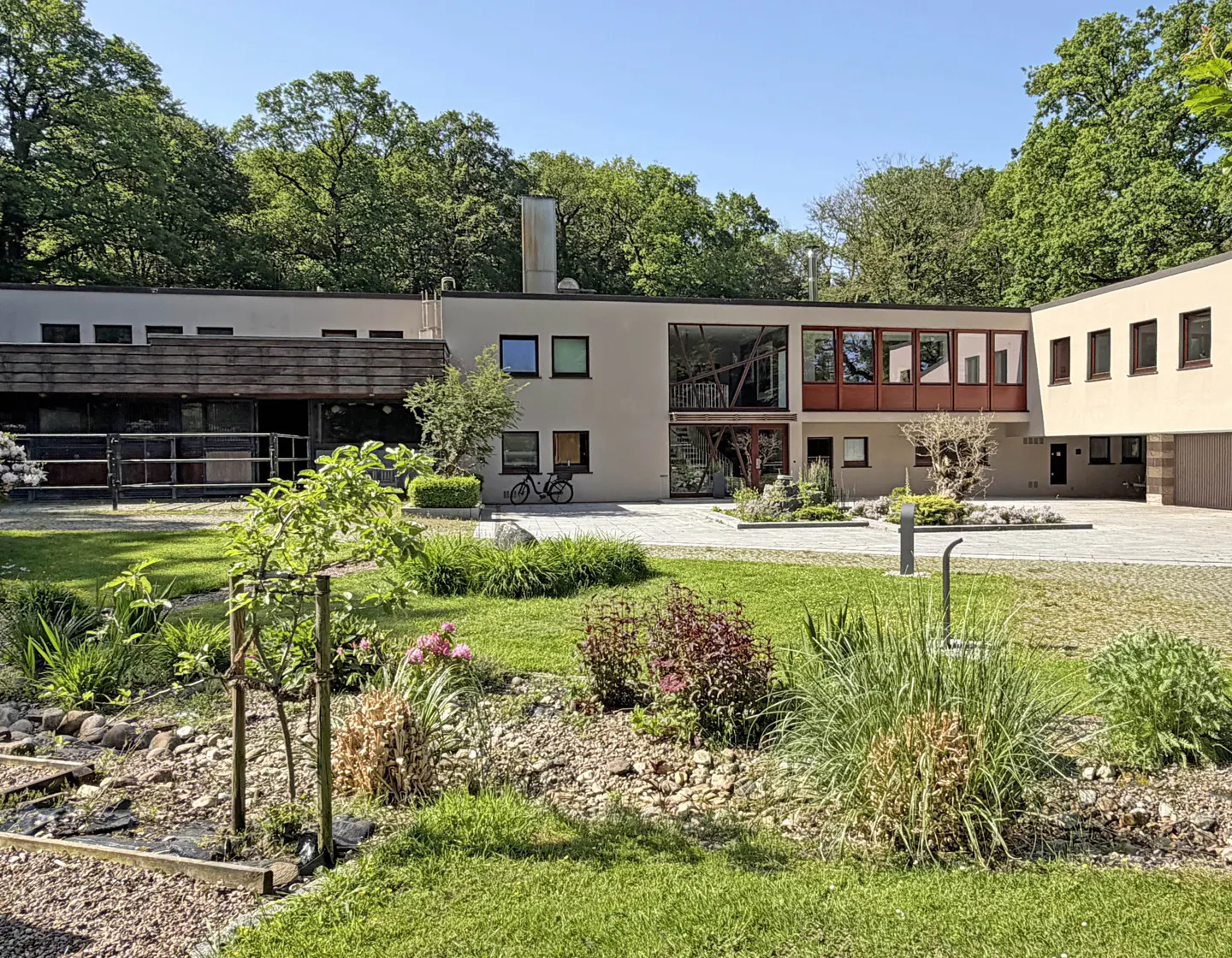 Modern beige building with a flat roof, large windows, and a landscaped front garden with shrubs and a paved driveway.