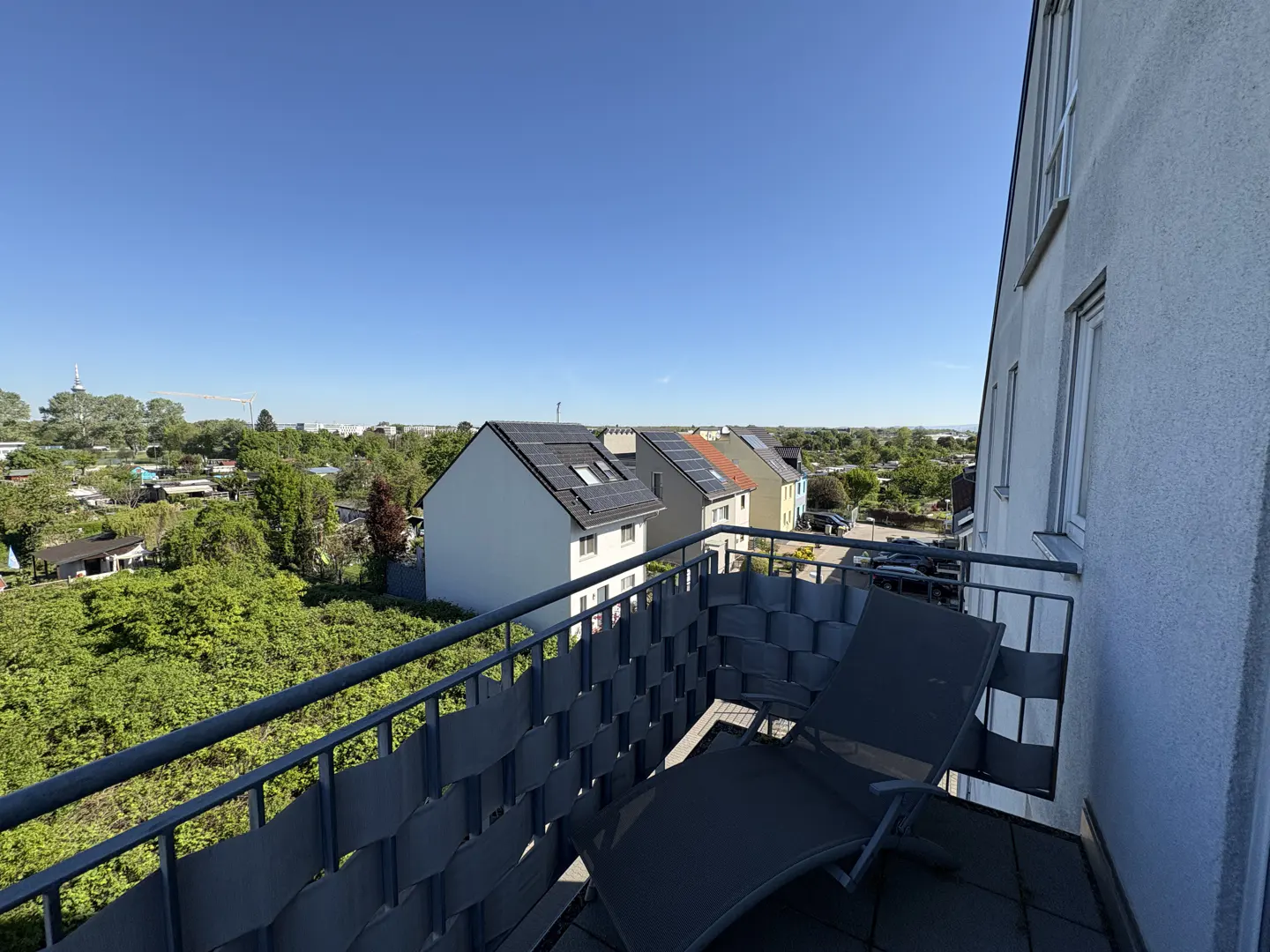 View from a balcony with a gray lounge chair, overlooking houses with solar panels and green trees under a clear blue sky.