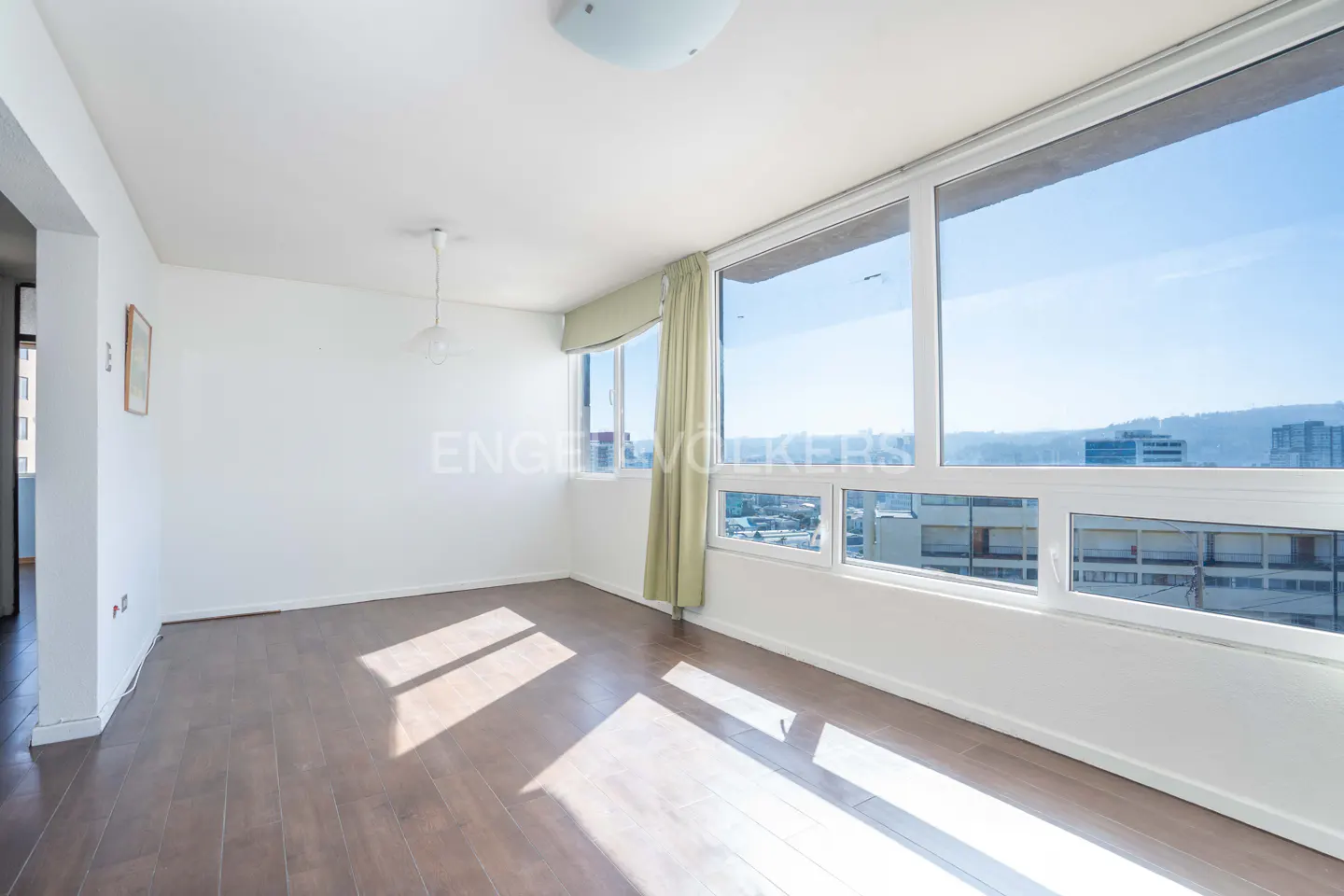 Bright, empty room with wood floors, white walls, and large windows showing a city view. A light green curtain hangs near the window.
