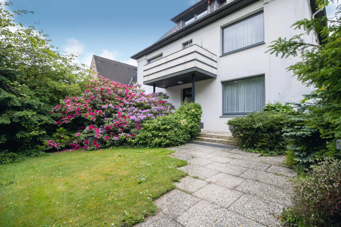 White house with a balcony, stone patio, and a lush garden featuring vibrant pink and purple rhododendrons.