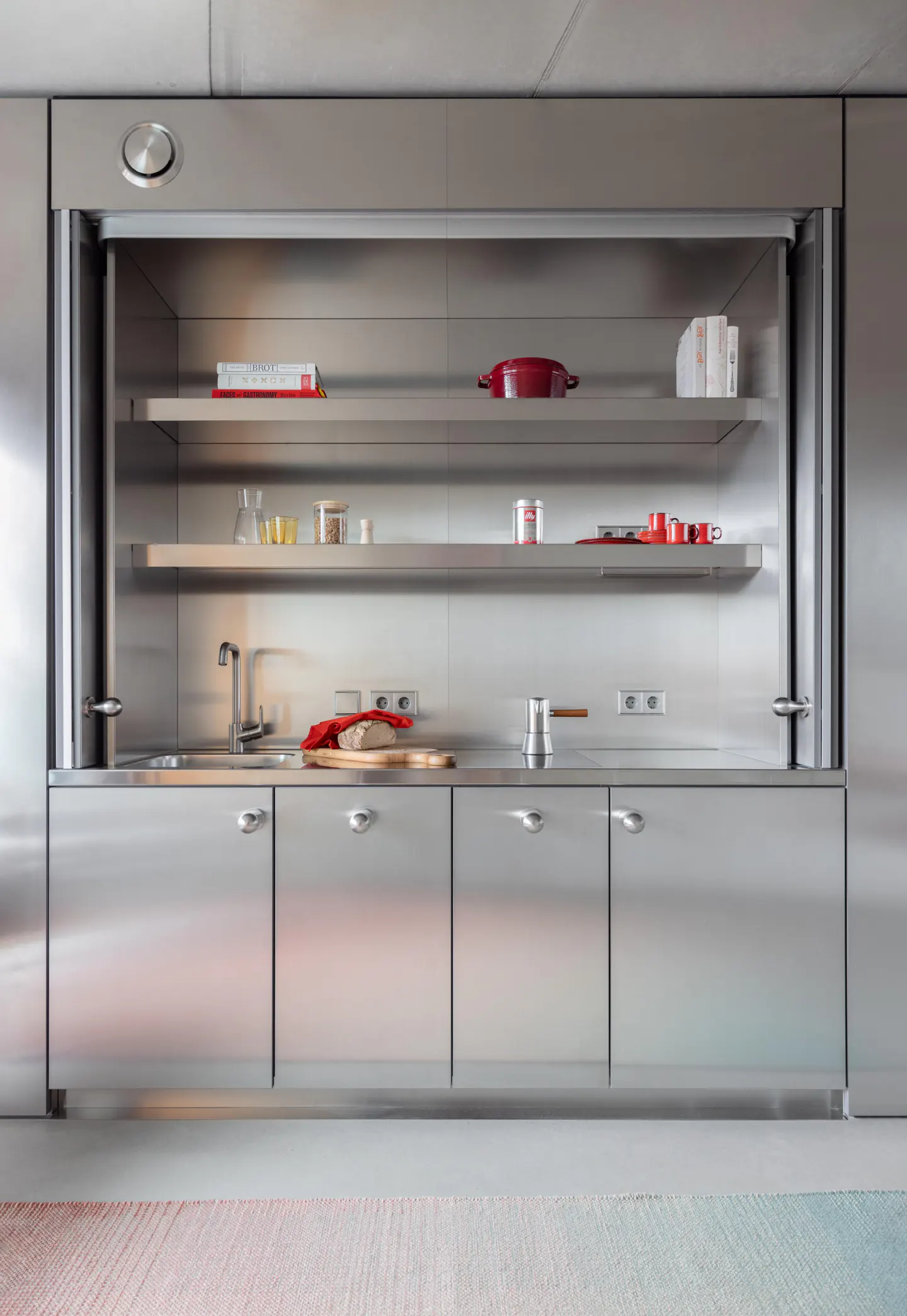 Modern kitchen with stainless steel cabinets, shelves, and sink. Red pot and dish towel add color.