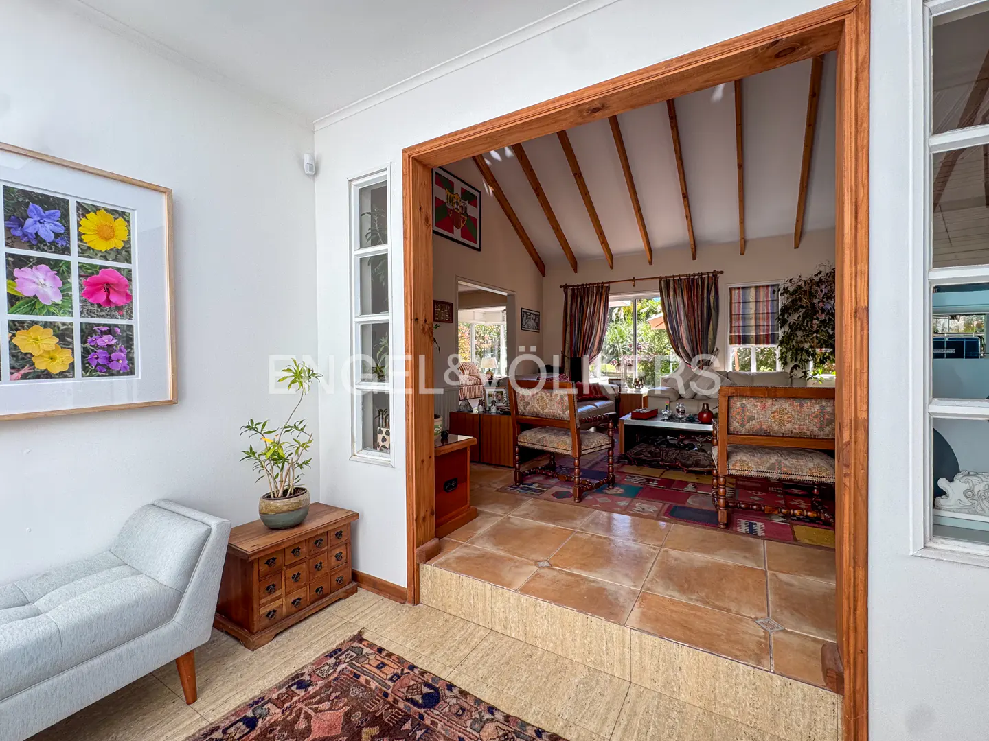 Interior view of a bright living room with a wooden frame doorway, colorful rug, and floral artwork on the wall.