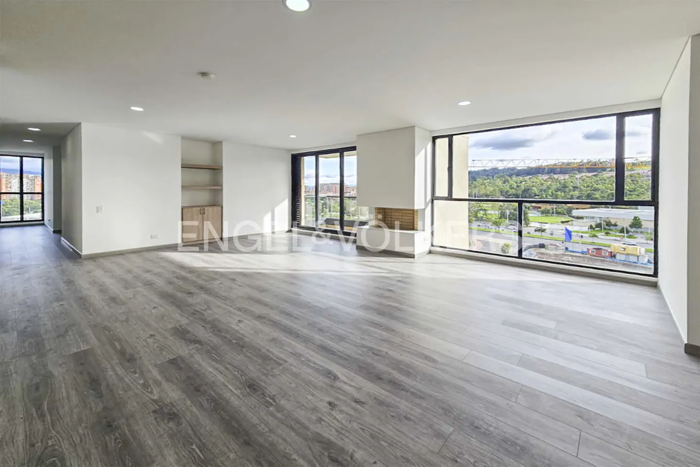 Spacious, empty living room with light gray wood floors, white walls, built-in shelving, and large windows with a city view.