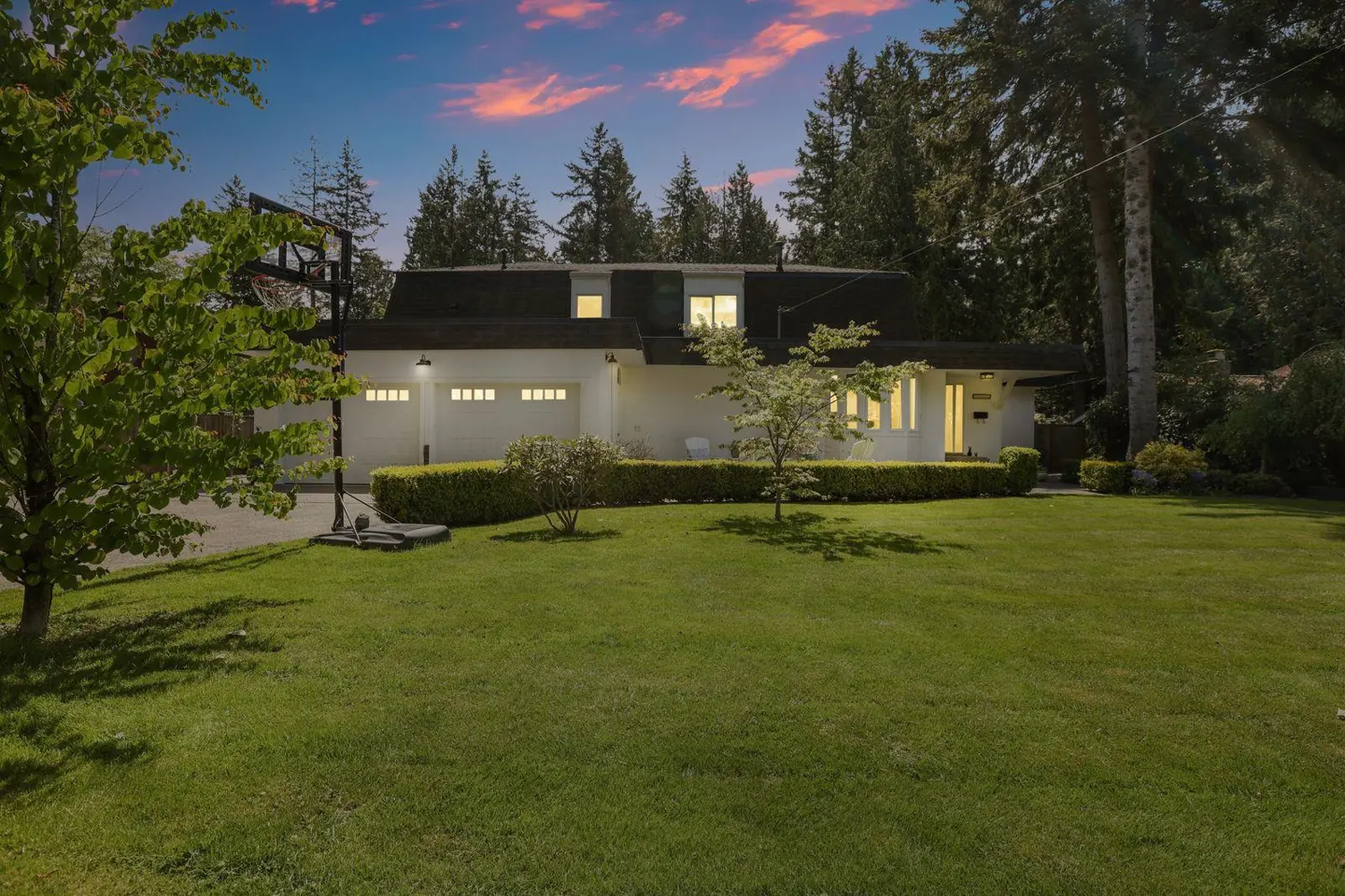 Exterior view of a one-story white house with a black roof, green lawn, and trees against a colorful sunset sky.