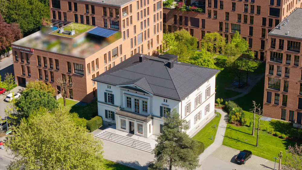 Aerial view of a white, two-story building with a gray roof, surrounded by brick buildings and green trees.