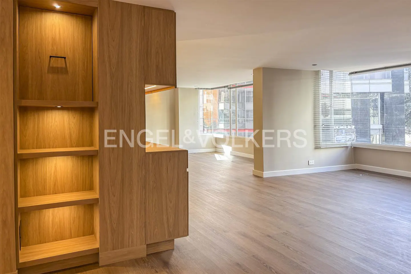 A bright, empty room with wood floors and a built-in wood shelving unit. Windows with blinds are visible in the background.