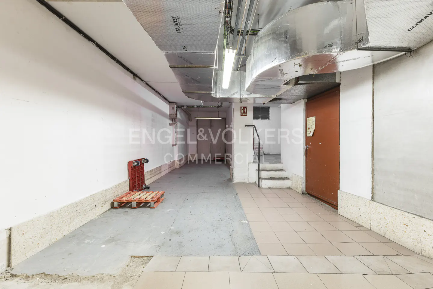 A commercial space with white walls, gray floors, and exposed ductwork on the ceiling. A red pallet and cart are on the left.