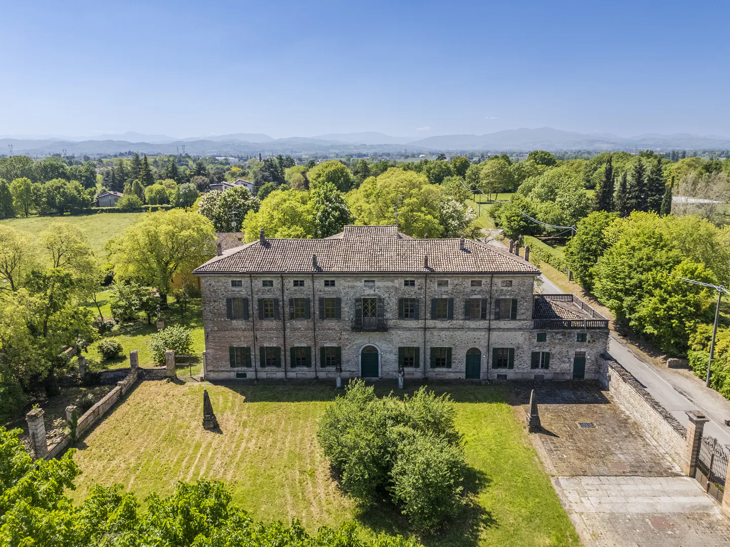 Aerial view of a large, two-story stone house with a red tile roof, green shutters, and a lawn surrounded by trees.