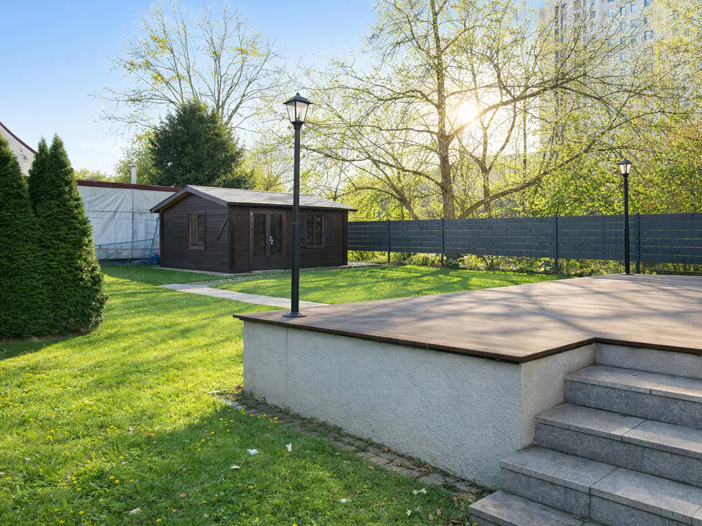 A backyard with a brown shed, green grass, trees, and a gray patio with steps.