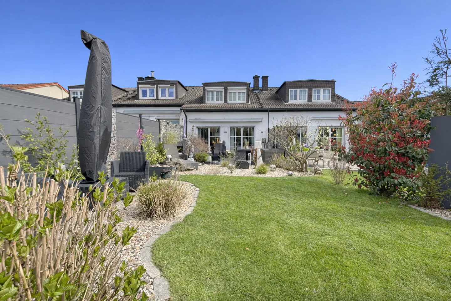 A well-manicured lawn leads to a white house with dormer windows. Patio furniture sits outside the sliding glass doors.