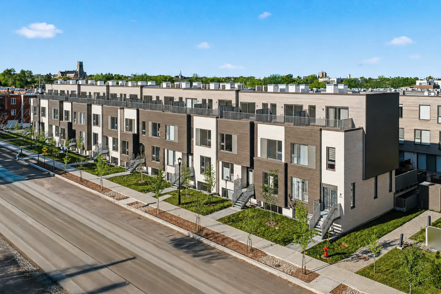 Row of modern townhouses with brown and white facades, small front yards, and rooftop terraces under a blue sky.