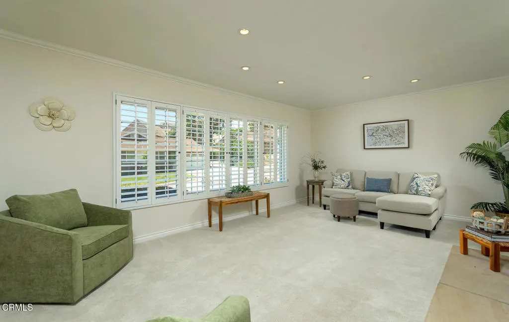 Living room with white walls, carpet, and shutters. Green armchair, beige sofa, and wooden table. Natural light.